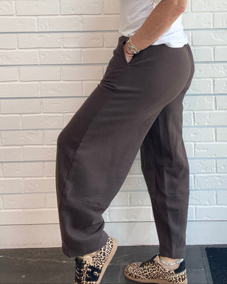 A woman wearing dark brown wide-leg jogger pants, a white tank top, and leopard print shoes, standing in front of a white brick wall.