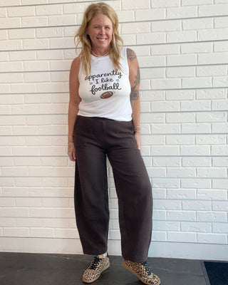 A woman stands against a white brick wall wearing a white graphic tank top that reads 'Apparently I like football', dark brown wide-leg pants, and leopard-print sneakers.