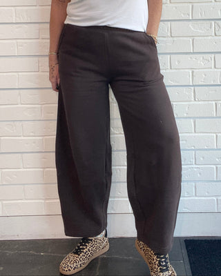 A woman stands against a white brick wall wearing a white sleeveless tank top with the text 'Apparently I like football' and a football graphic, paired with dark wide-leg pants and leopard-print shoes.