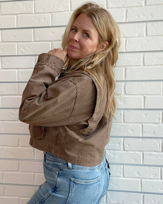 A woman with blonde hair stands against a white brick wall, wearing a brown cropped utility jacket and light blue jeans.