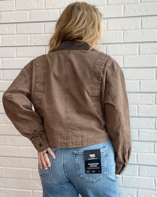 A woman viewed from behind wearing a brown cropped utility jacket and light blue jeans, standing against a white painted brick wall. A black NIK brand label is visible on the back pocket of the jeans.