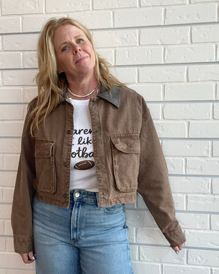 A woman wearing a brown cropped utility jacket over a white graphic t-shirt and blue jeans, standing against a white brick wall.