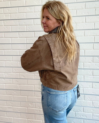 A woman with blonde hair is shown from the back wearing a brown cropped utility jacket and light blue jeans, standing against a white brick wall.