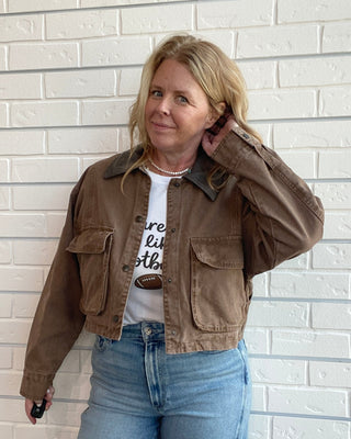 A woman wearing a brown cropped utility jacket over a white graphic t-shirt and light blue jeans, posing against a white brick wall.