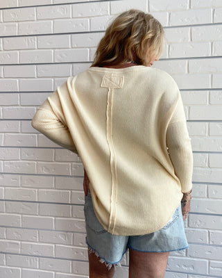 A woman viewed from behind wearing a cream-colored knit sweater and denim shorts, standing against a white brick wall.