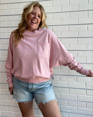 A woman wearing a dusty pink long-sleeve crewneck sweatshirt with a chest pocket and light blue denim shorts, posing against a white brick wall.