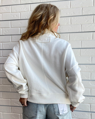 A woman viewed from behind wearing an off-white/cream crewneck sweatshirt and light-wash denim shorts, standing against a white painted brick wall.