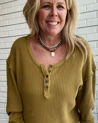 A smiling blonde woman wearing a textured mustard yellow waffle-knit henley sweater with a button placket, standing against a white brick wall.