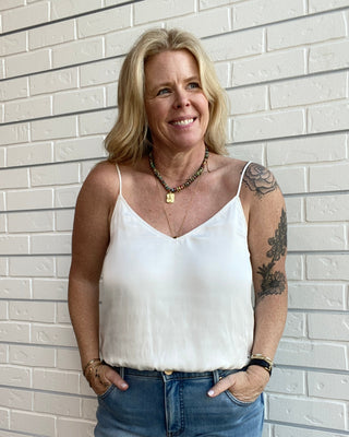 Woman wearing a white top and blue jeans standing against a light brick wall.