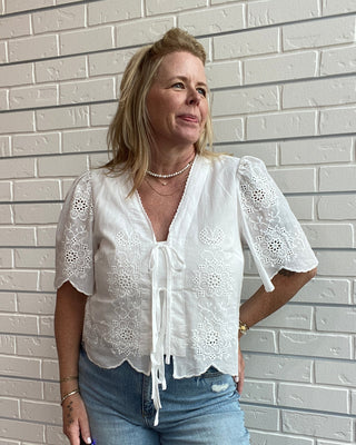 Woman wearing a white blouse with lace details and blue jeans against a light tiled wall.