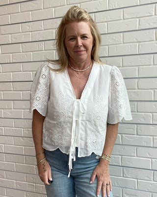 Woman wearing a white blouse and blue jeans standing against a light brick wall.