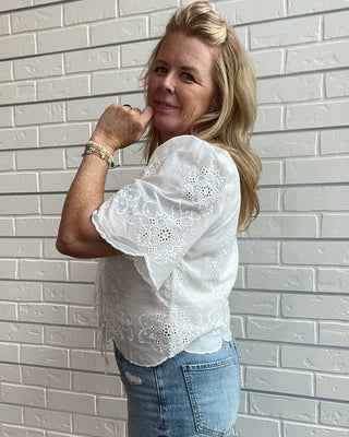Person wearing a white blouse with lace details and blue jeans against a light gray tiled wall.