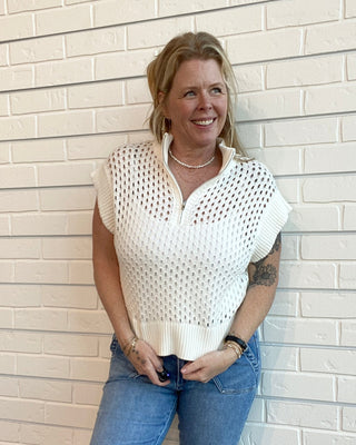 Woman wearing a white open-knit top and blue jeans standing against a light brick wall.