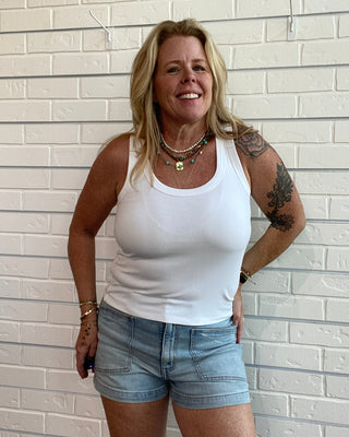A smiling blonde woman with tattoos stands against a white brick wall, wearing a white tank top and light blue denim shorts.