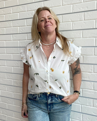 Woman wearing a white blouse with floral patterns and blue jeans against a white brick wall.