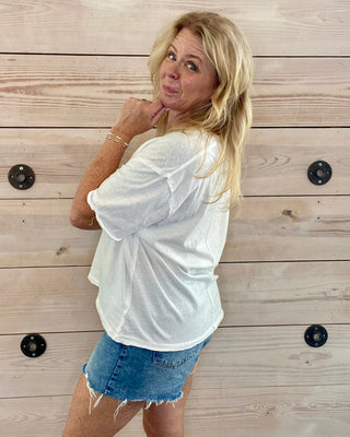 Woman wearing a white shirt and denim shorts standing against a wooden wall.
