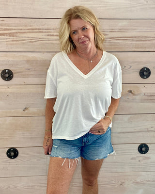 Woman wearing a white t-shirt and denim shorts standing against a wooden wall.
