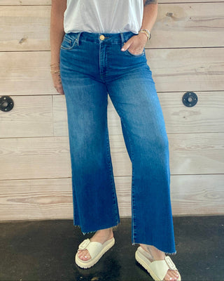 Person wearing a white t-shirt and blue jeans standing against a wooden wall.