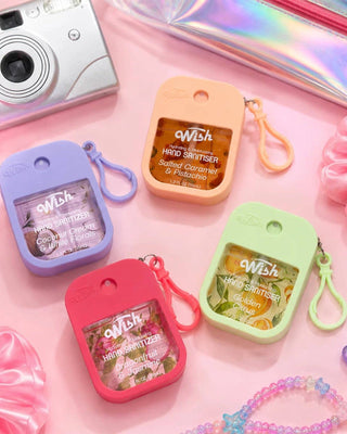 Four small Wish brand hand sanitizer pouches in pastel purple, peach, pink, and light green, arranged on a pink surface with a silver camera, iridescent pouch, scrunchies, and beaded accessories around them.