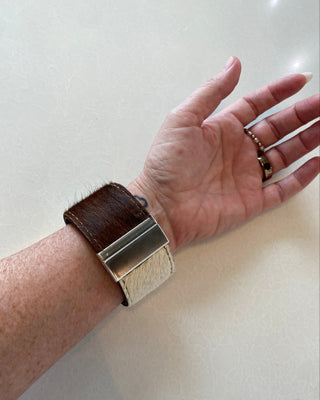 A close-up of a person's wrist wearing a unique watch with a brown and white textured strap and a large silver rectangular clasp, set against a light beige background.