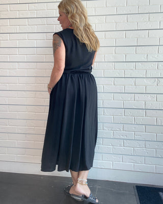 A woman with blonde hair is shown from the back wearing a sleeveless black midi skirt top set with side pockets, standing against a white brick wall.