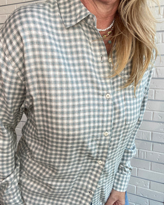 A woman wearing a long-sleeve button-up shirt with a muted green and beige gingham pattern, standing against a white painted brick wall.