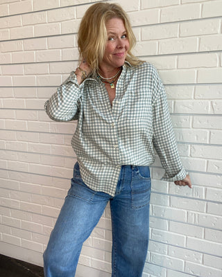 A woman wearing a light sage green and white gingham button-down shirt with blue jeans, standing against a white brick wall.