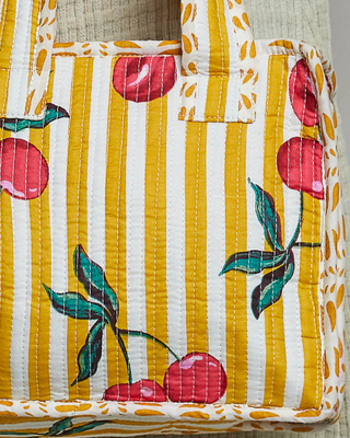 A quilted fabric tote bag featuring a yellow and white vertical stripe pattern with red cherries and green leaves printed across the front, set against a neutral textured background.