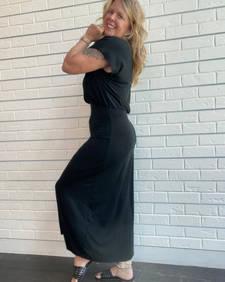 A woman modeling a black short-sleeve maxi dress with a draped, gathered bodice and side slit, standing against a white painted brick wall.
