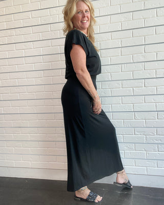 A woman modeling a black short-sleeve maxi dress with a draped neckline, standing against a white brick wall.