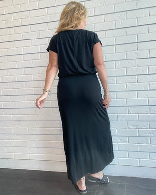 A woman viewed from behind wearing a black short-sleeve top and a long black maxi skirt, standing against a white painted brick wall.
