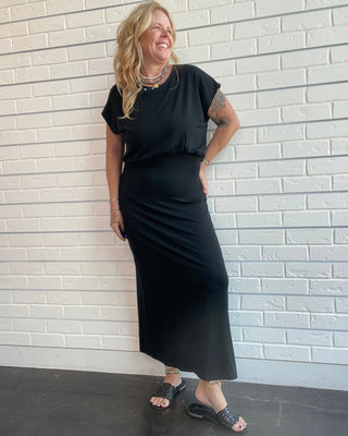 A woman wearing a black short-sleeve top and matching black midi skirt, standing against a white brick wall.