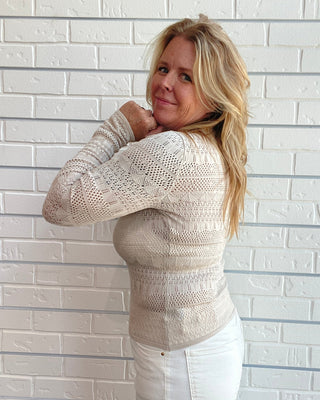 A woman with blonde hair wearing a cream-colored, textured knit sweater and white pants, posing against a white brick wall.