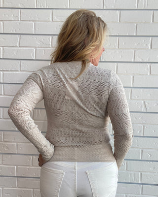 A woman viewed from behind wearing a beige open-knit long-sleeve sweater and white pants, standing against a white brick wall.