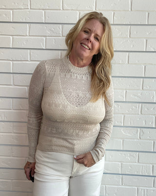 A smiling blonde woman wearing a beige knit mock-neck sweater with a lace overlay and white pants, standing against a white brick wall.