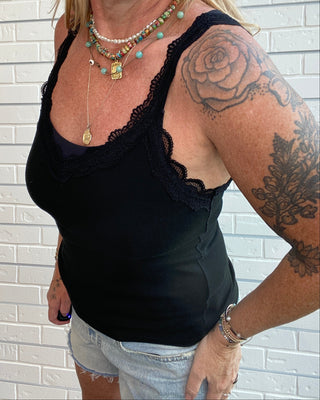 A woman wearing a black lace-trimmed camisole tank top and light-wash denim shorts, standing against a white brick wall. She is wearing layered necklaces and has a large rose tattoo on her right arm.
