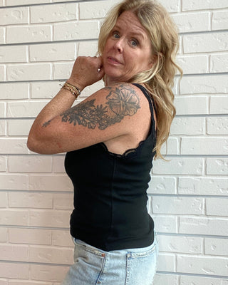 A woman with blonde hair and a visible tattoo on her upper arm poses against a white brick wall. She is wearing a black sleeveless top and a light-wash denim mini skirt.