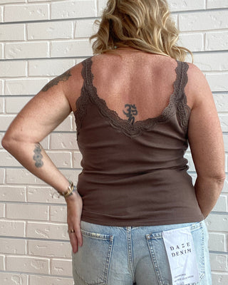 A woman shown from behind wearing a brown lace-trimmed tank top and light-wash denim shorts, standing against a white brick wall. A paper tag on the back of the shorts reads 'DAZE DAZE'.