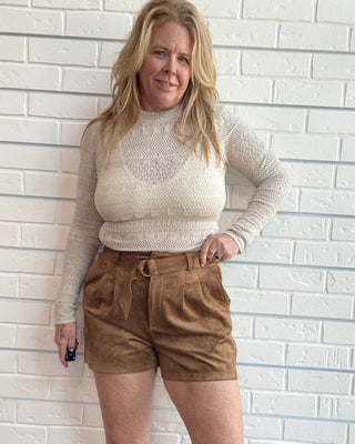 A woman stands against a white brick wall wearing a cream knit mock-neck top and brown corduroy shorts, paired with beige sandals.