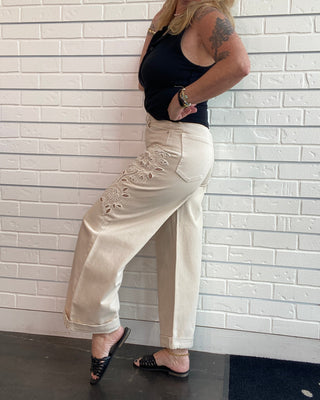 A woman wearing cream wide-leg pants with floral embroidery on the side and a black tank top, posing against a white brick wall.