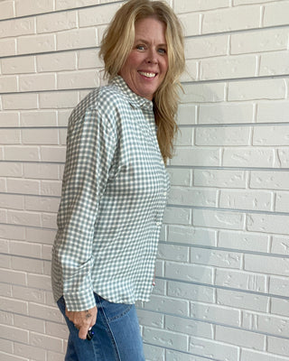 A woman with blonde hair smiling and posing in a light green and white gingham long-sleeve button-down shirt with blue jeans, standing against a white brick wall.