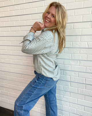 A woman with blonde hair wearing a light green and white gingham long-sleeve button-up shirt and blue jeans, posing against a white brick wall.