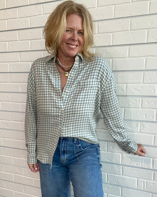 A woman with blonde hair stands against a white brick wall wearing a light sage green and white gingham button-down shirt and blue jeans.