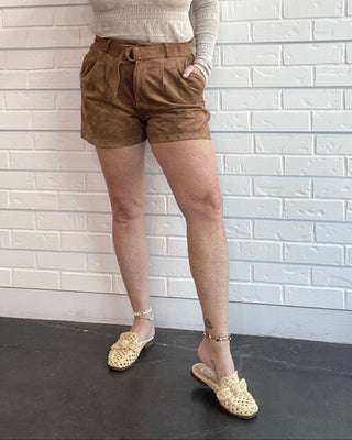 A woman modeling a cream knit mock-neck sweater and brown corduroy shorts, paired with gold woven mule sandals, standing against a white brick wall.