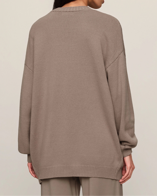 A woman shown from the back wearing an oversized knit sweater in a taupe-brown color, paired with matching pants, against a plain light beige studio background.
