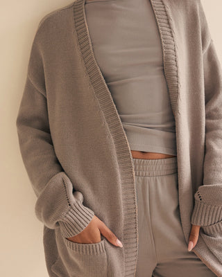 A person wearing a taupe open-front knit cardigan over a matching ribbed top and jogger pants, photographed against a plain warm beige background.