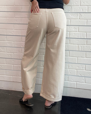 A woman wearing beige wide-leg cropped pants and a black sleeveless top, shown from a side/back angle against a white brick wall.