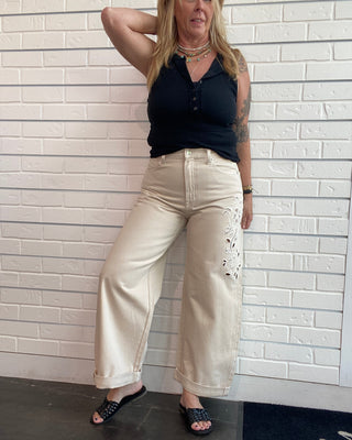 A woman wearing beige wide-leg cropped pants with white embroidery on the thigh, a black sleeveless top, and black slide sandals, posing against a white brick wall.