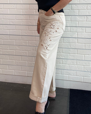 A woman wearing off-white cropped pants with floral embroidery on the thigh and a black sleeveless top, posing against a white brick wall.