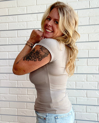 A woman with blonde hair and a visible tattoo on her right arm poses in front of a white brick wall. She is wearing a fitted beige short-sleeve top and a light blue denim mini skirt.